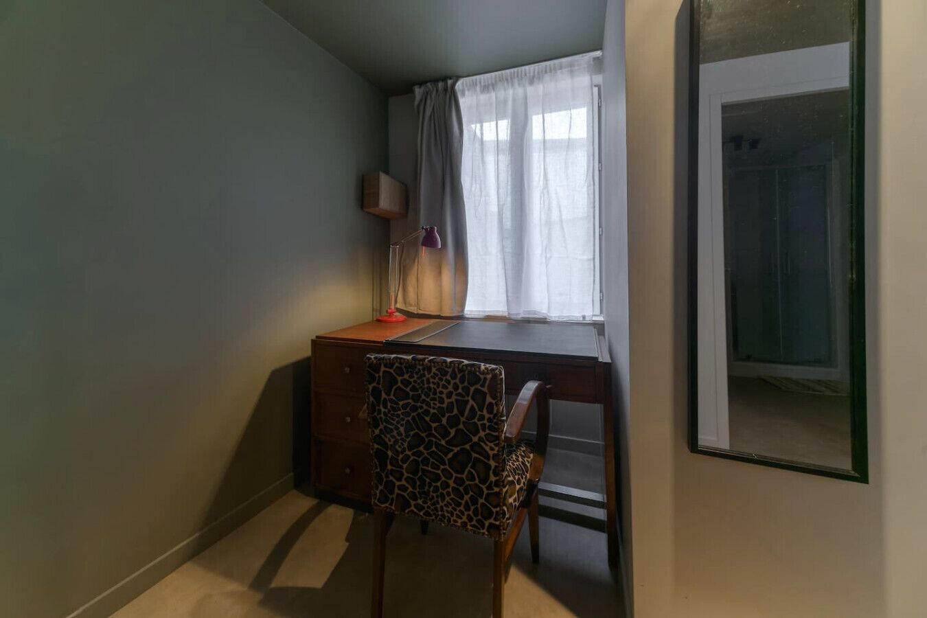 Ein kleiner, schummrig beleuchteter Raum mit Schreibtisch, Stuhl, Lampe und einem Fenster mit durchsichtigen Vorhängen.