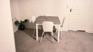A small, sparsely furnished room with a single dining table and four chairs. A door is visible on the right, and a painting hangs on the wall above the table. - Appartement Cosy en centre ville (Clermont-Ferrand)