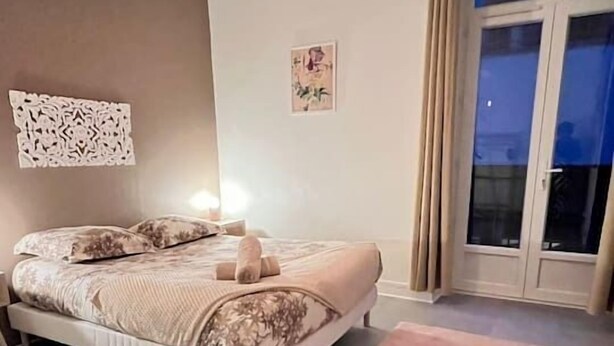 A bedroom with a bed, a bedside table, a hanging light fixture, a window with curtains, and a view of the outside. - Appartement Cosy en centre ville (Clermont-Ferrand)