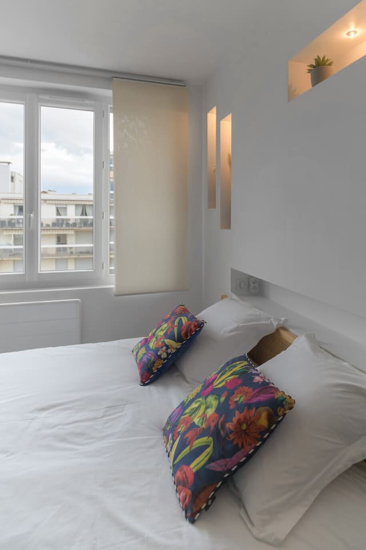 Habitación de hotel con una cama grande, dos almohadas coloridas, una mesita de noche, una pequeña planta en maceta y una ventana con vistas a edificios.