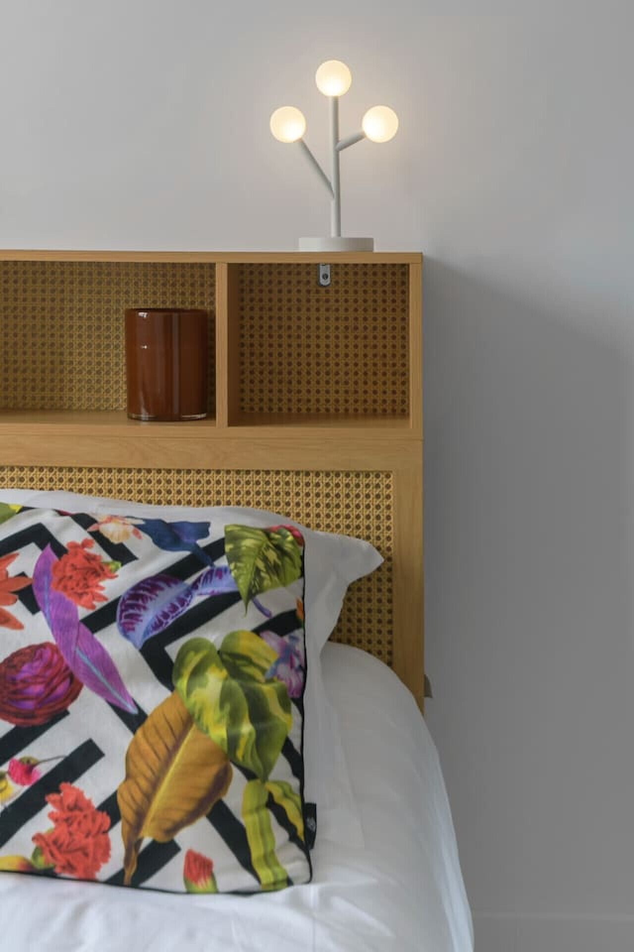 Una cama con un edredón floral colorido, un cabecero de madera con una lámpara y un pequeño florero marrón en la estantería.