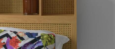 Una cama con un edredón floral colorido, un cabecero de madera con una lámpara y un pequeño florero marrón en la estantería.
