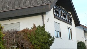 Um edifício de dois andares com telhado inclinado, varanda no andar superior e um pequeno jardim frontal com uma árvore e arbustos.