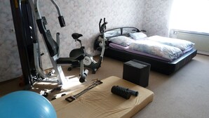 Quarto com cama, equipamentos de ginástica, uma bola de exercício azul e uma plataforma de madeira com halteres.
