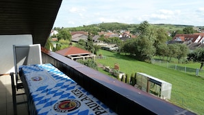 Uma varanda com uma mesa coberta por um tecido xadrez, vista para uma paisagem verde com casas e um céu claro.