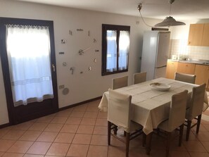 Ein Essbereich mit einem für vier Personen gedecktem Tisch, ein Küchenbereich mit einem Kühlschrank und ein Fenster mit Vorhängen.