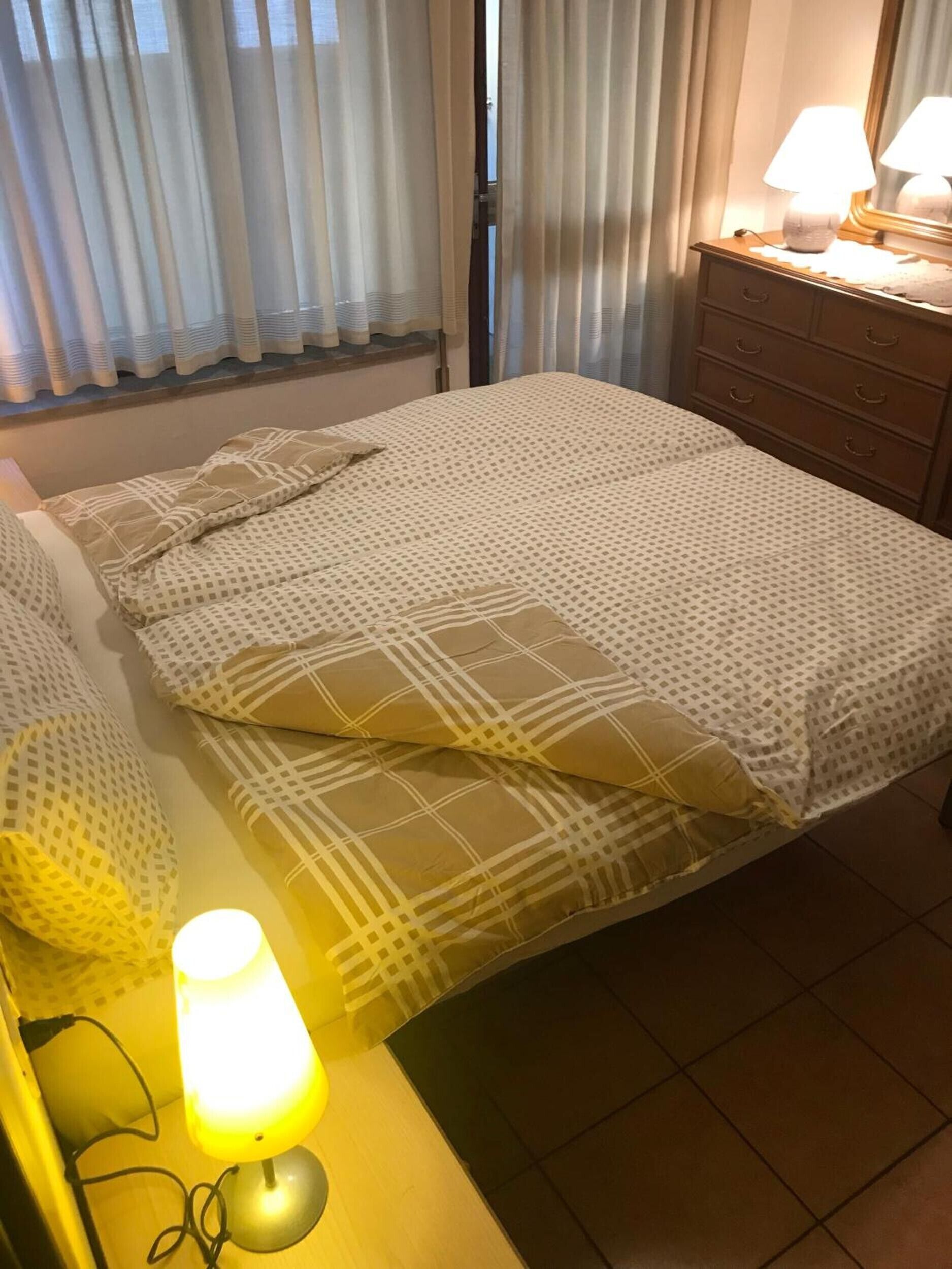Un letto rifatto con biancheria a motivi, una lampada da comodino e una cassettiera con specchio.