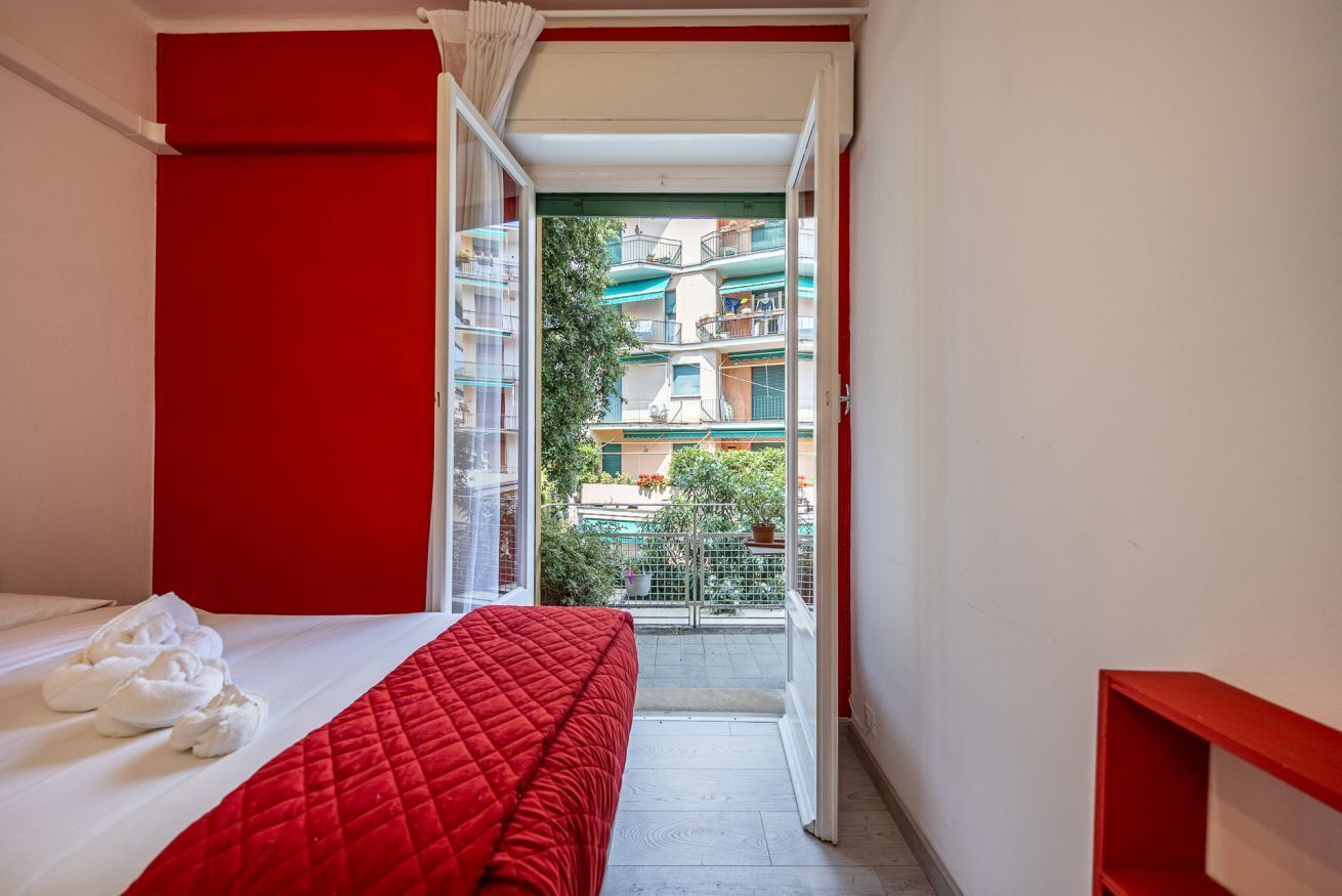 A narrow hotel room with a red bedspread, a white bed, and a view of a balcony with buildings.