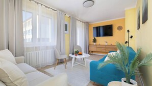 A modern living room with a sofa, a blue armchair, a small round table with a plant, and a TV mounted on the wall. - Colorful Apartment in Wisla | 2 Bedrooms | Workspace. (Wisła)