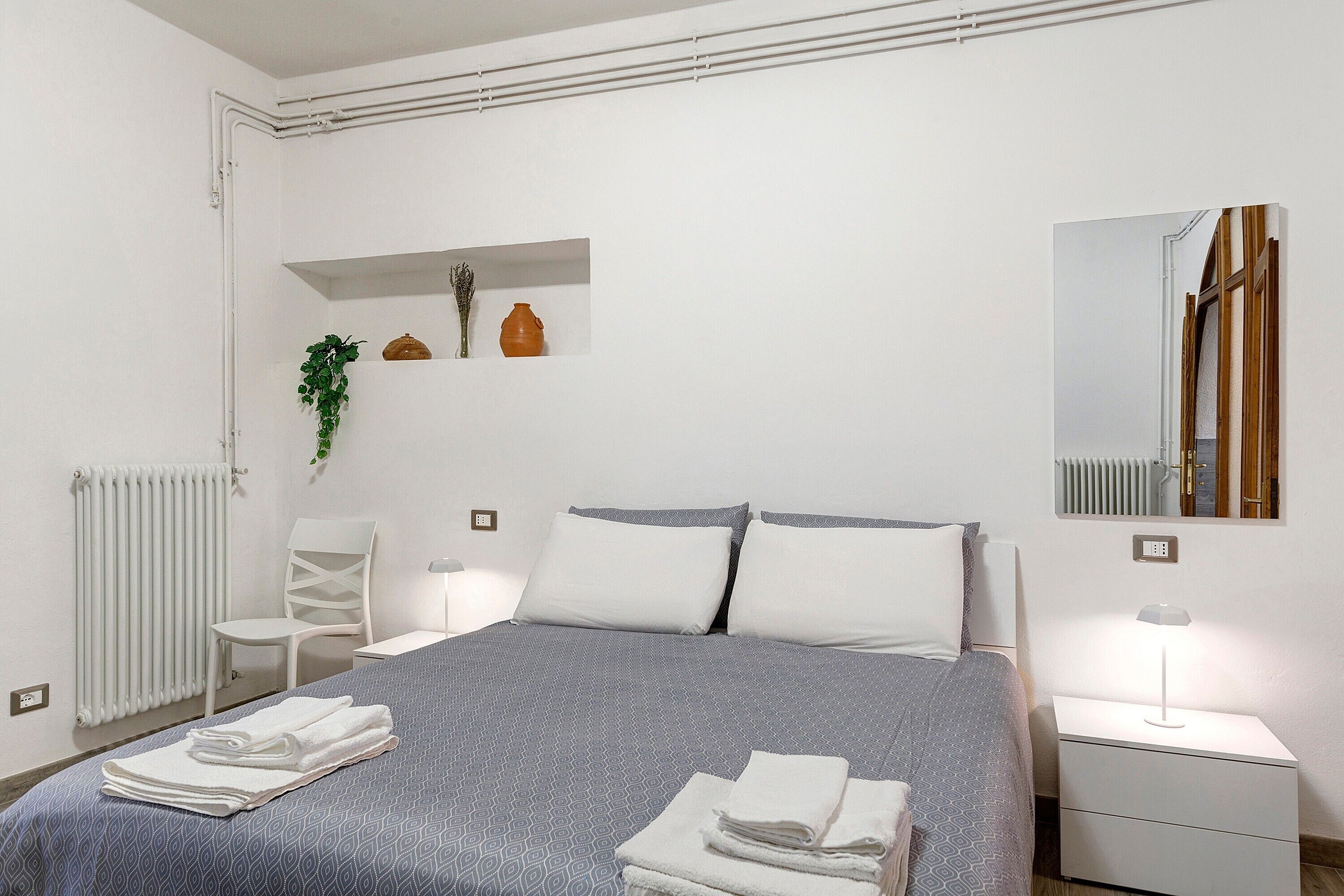 Ein ordentlich eingerichtetes Schlafzimmer mit Bett, Nachttischen, einem Sessel, einem Heizkörper und einem Regal mit Dekoration.