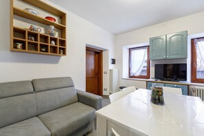 A living room with a grey sofa, a dining table with a clear glass vase, wooden shelves with decorative items, a wooden door, and a kitchen area with cabinets and a TV. - Apartment 'In Casetta Da Mae' with Mountain View (Lorenzago di Cadore)
