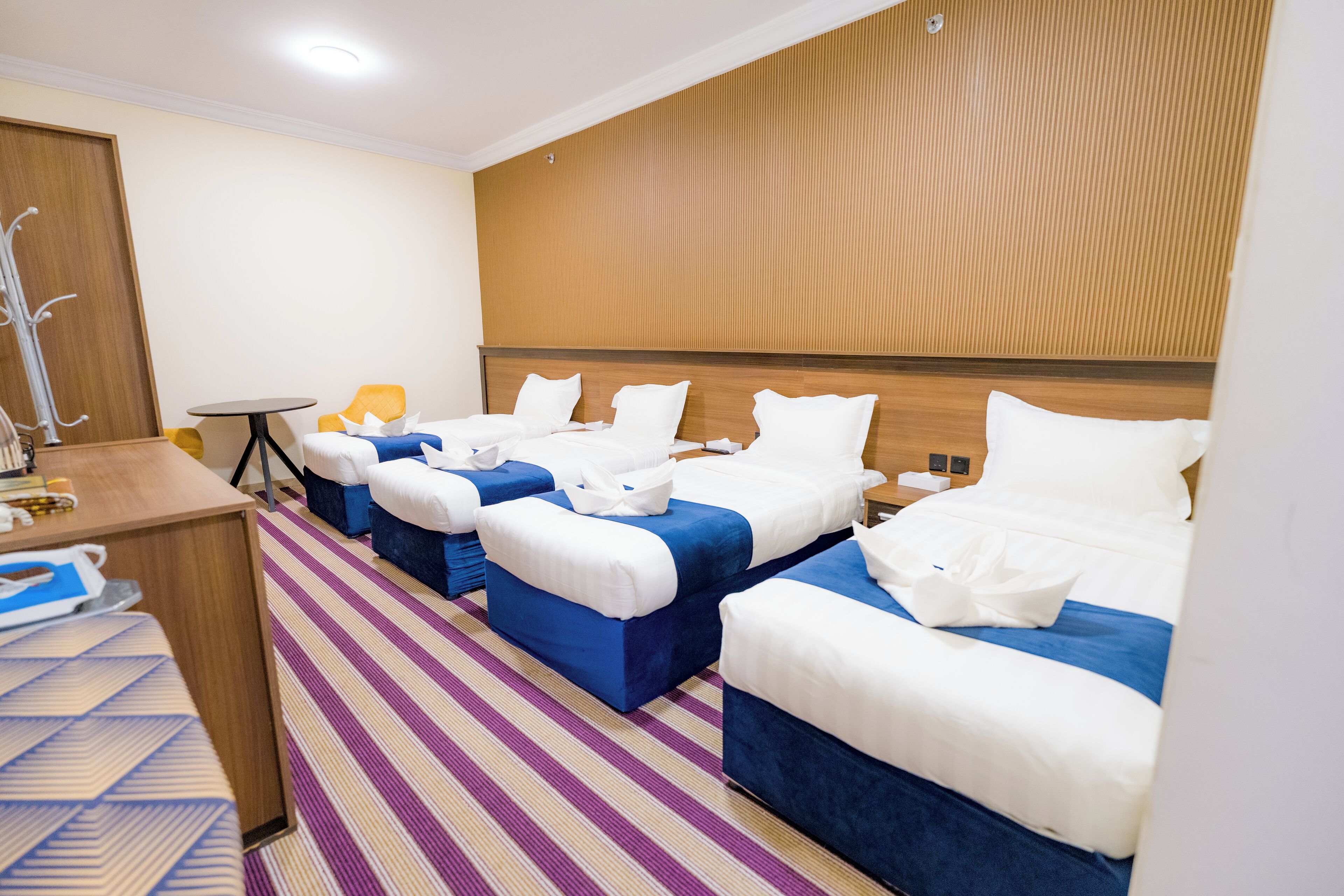 A hotel room with four beds arranged in a row, a small table, and a striped carpet.