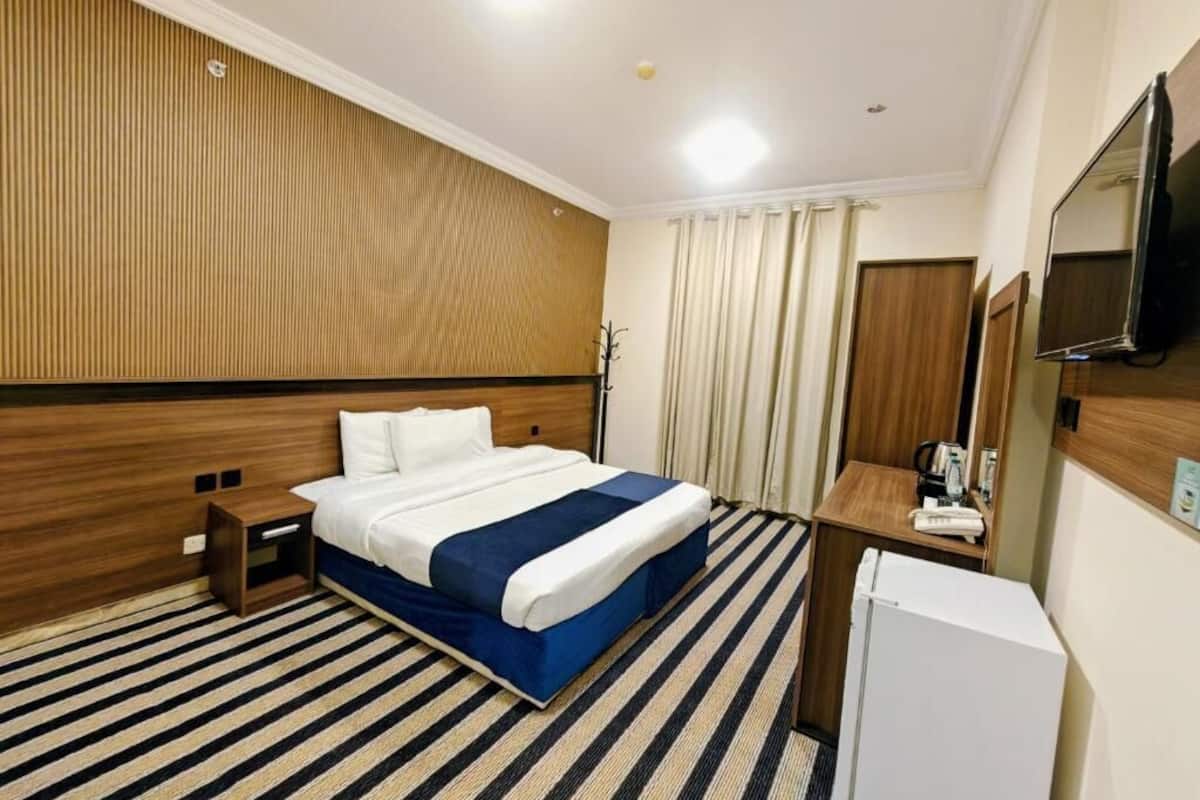 Habitación de hotel con cama, mesita de noche, escritorio pequeño con silla, televisión y refrigerador.