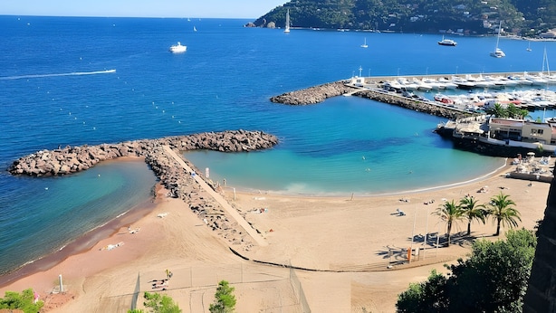 Eine Küstenansicht mit einem steinernen Wellenbrecher, einem Sandstrand und einem Jachthafen mit Booten.