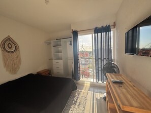 Une chambre à coucher comprenant un lit, un bureau en bois, une télévision, une armoire et un balcon avec des rideaux.