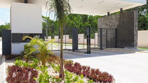 Un edificio moderno con entrada protegida por una reja, un paseo techado y jardines con diferentes plantas.