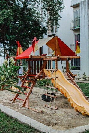 Un terrain de jeux avec un toboggan et des balançoires, entouré d’un bâtiment et de verdure.