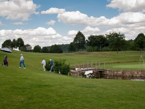 Ein Golfplatz mit einer Stegbrücke über einem kleinen Teich, umgeben von Bäumen und einem wolkenlosen Himmel.