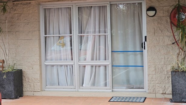 A building entrance with a stone facade, a sliding glass door with white curtains, and two potted plants on either side.