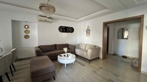 A living room with a brown sofa, a white coffee table, and a beige sofa. There is a chandelier hanging from the ceiling, and a clock on the wall. - Luxury apartment 170 m² La Marsa - 3 bedrooms, terrace & private parking (La Marsa)