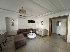 A living room with a brown sofa, a white coffee table, and a beige sofa. There is a chandelier hanging from the ceiling, and a clock on the wall. - Luxury apartment 170 m² La Marsa - 3 bedrooms, terrace & private parking (La Marsa)