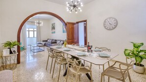 A dining area with a long table set for six, surrounded by chairs, a chandelier, and a clock on the wall. - Traditional, large 3 bedroom house with Yard JBAR1-1 (St Julian's)