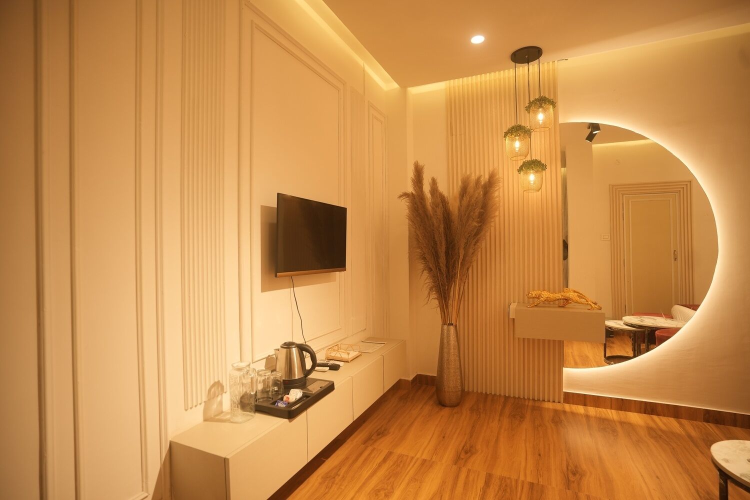 Una camera d'albergo moderna con pavimento in legno, televisore a schermo piatto montato a parete, un piccolo tavolino consolle con bollitore e bicchieri, e un grande specchio con cornice circolare.