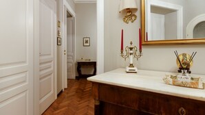 Een hal met witte deuren, een houten vloer, een witte dressoir met spiegel, een kandelaar en een kleine tafel met een lamp.