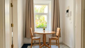 Una habitación con una mesa redonda de madera, dos sillas de madera, una ventana con cortinas, un radiador y un teléfono montado en la pared.