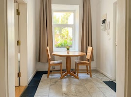 A room with a round wooden table, two wooden chairs, a window with curtains, a radiator, and a wall-mounted phone.