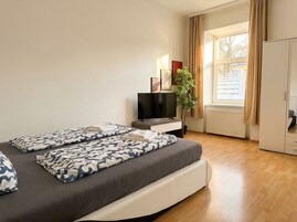 A hotel room with a bed, a TV, a plant, and a window with curtains.