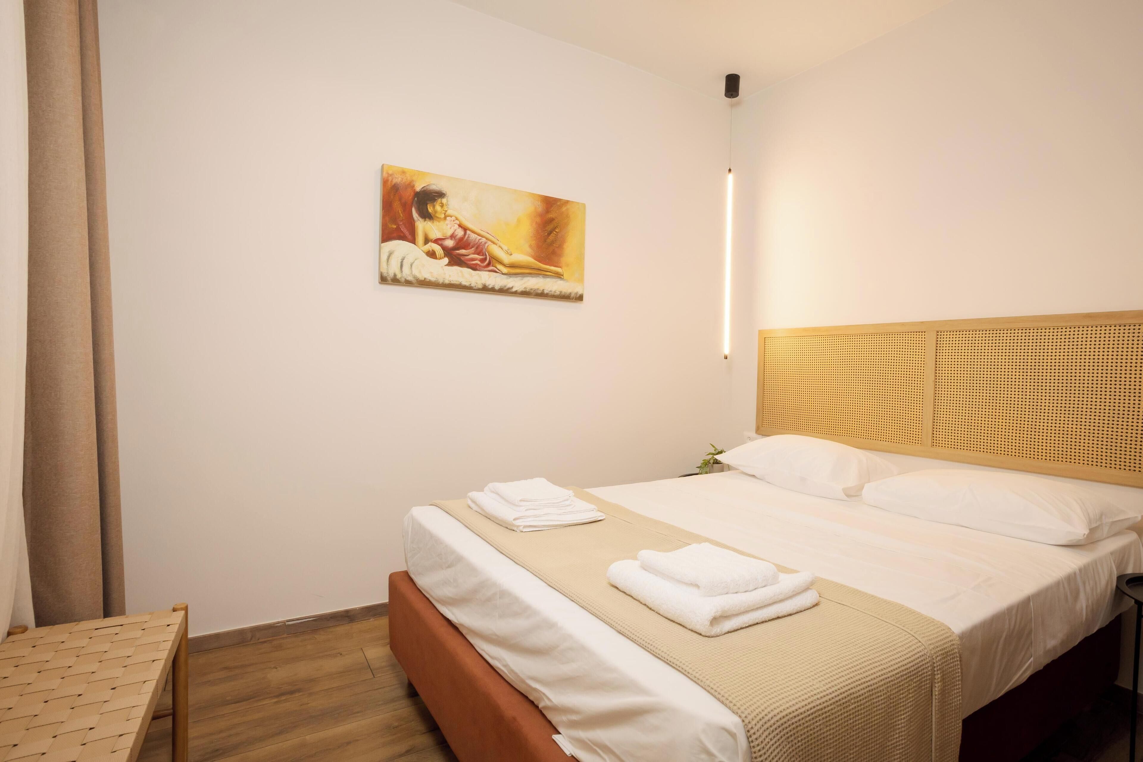A neatly made bed with white linens and a beige blanket, a painting on the wall, and a wooden headboard.