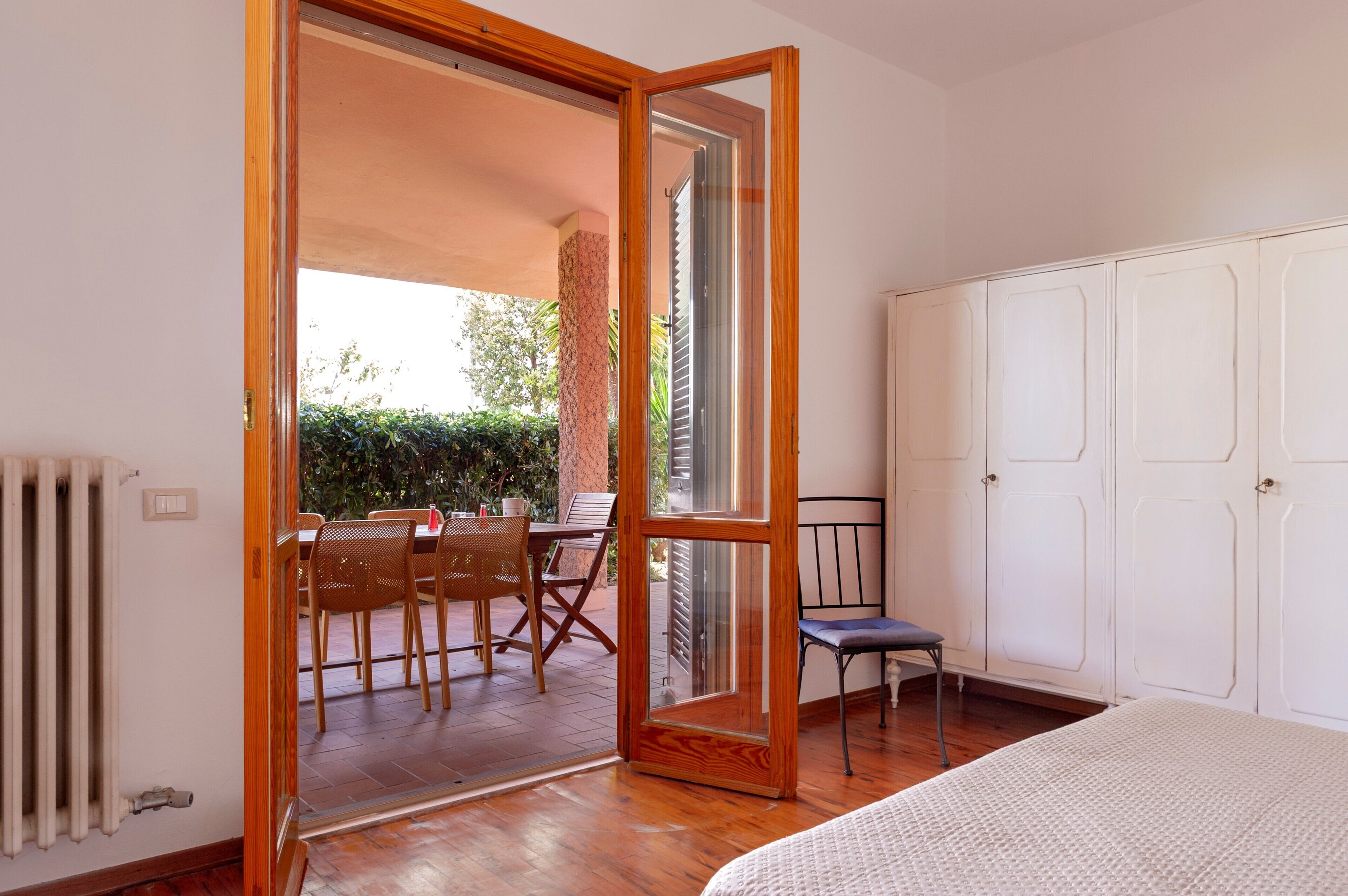 Une pièce dotée d’un plancher en bois, d’un lit, d’une armoire blanche et d’une porte-fenêtre coulissante donnant sur une terrasse extérieure avec une table et des chaises.