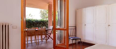 Une pièce dotée d’un plancher en bois, d’un lit, d’une armoire blanche et d’une porte-fenêtre coulissante donnant sur une terrasse extérieure avec une table et des chaises.