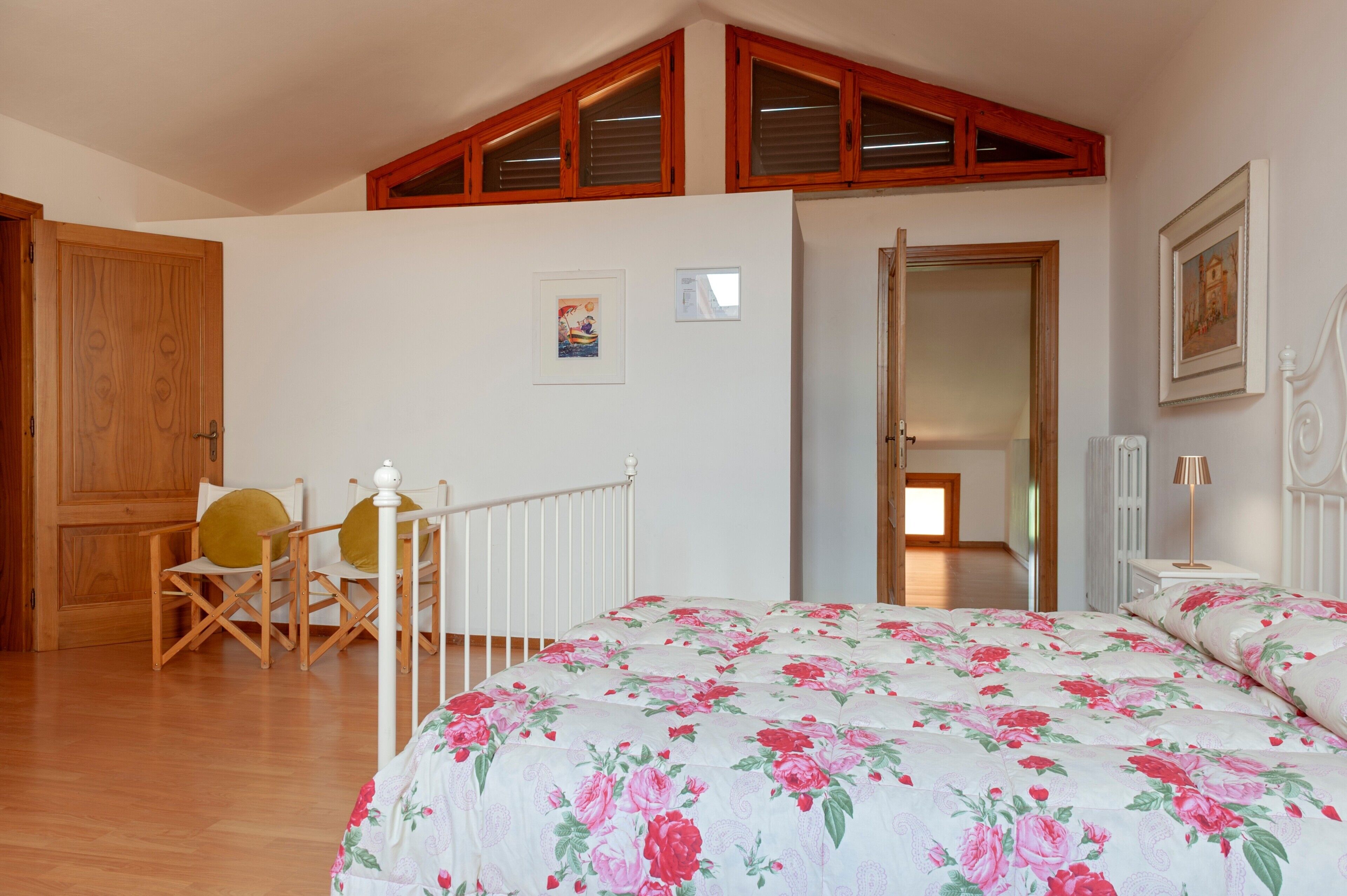 Une chambre avec un plafond en bois, un lit recouvert d’une literie à motifs floraux, une porte en bois, une chaise et une table de chevet avec une lampe.