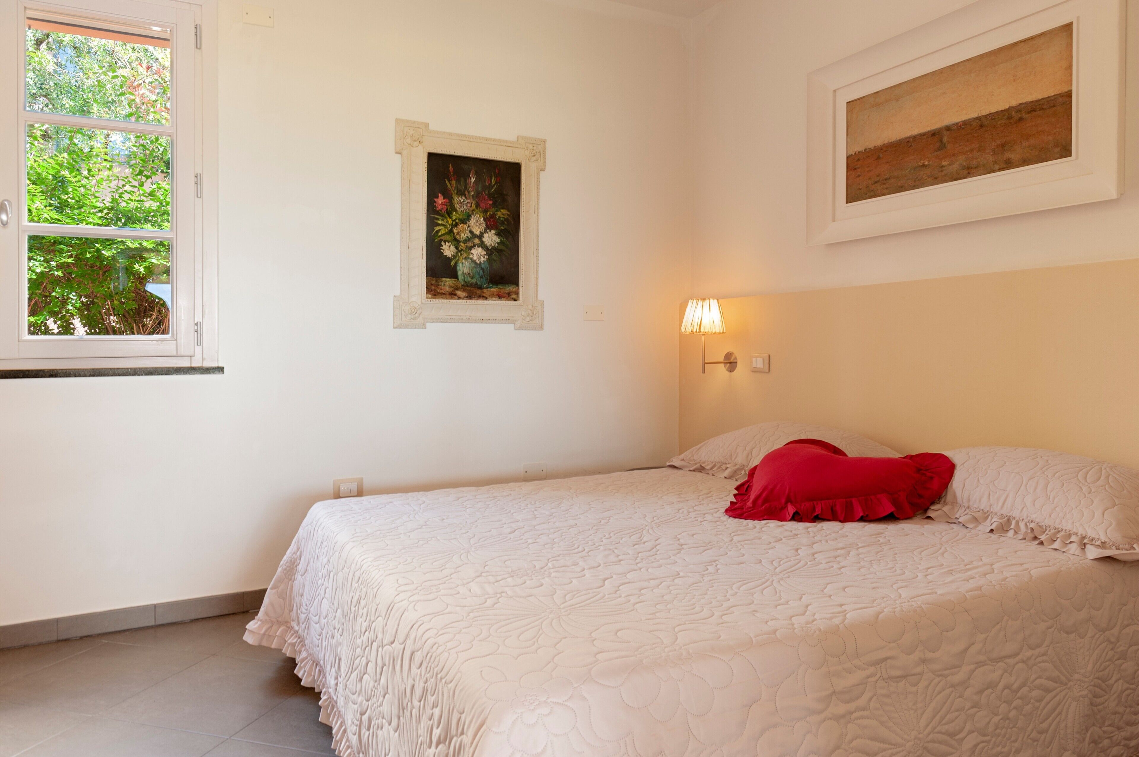 Ein Schlafzimmer mit einem Bett, einem Fenster, einer Wandlampe und zwei gerahmten Bildern.