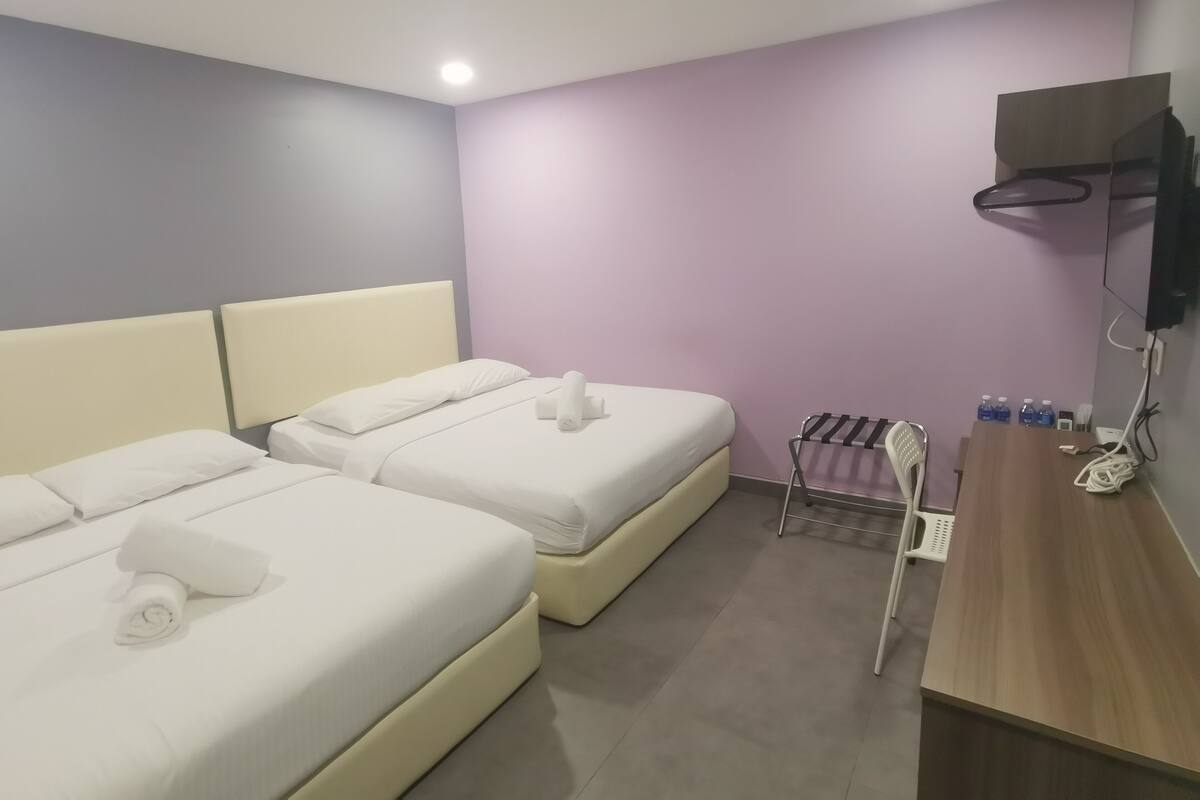 Ein Hotelzimmer mit zwei Betten, einem Holztisch, einem an der Wand montierten Fernseher und einer kleinen Sitzecke mit einem Sessel und einem Flaschenregal.