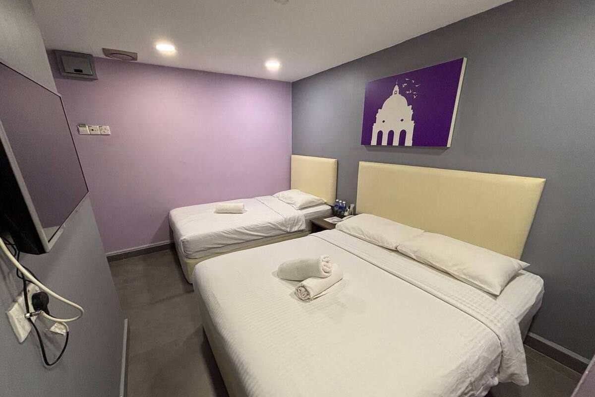 Ein Hotelzimmer mit zwei Betten, jeweils mit Kopfteil und Kissen. An der Wand ist ein Fernseher befestigt und ein Gemälde eines Gebäudes hängt dort.
