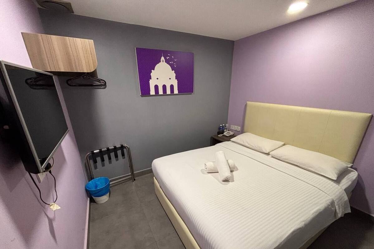 Ein Hotelzimmer mit einem Bett, einem Schreibtisch mit Computer, einem Wandfernseher, einem Gemälde und einer kleinen Mülleimer.