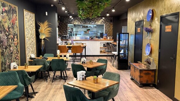 En restauranginteriör med grönt klädda stolar, träbord och ett centralt köksområde.