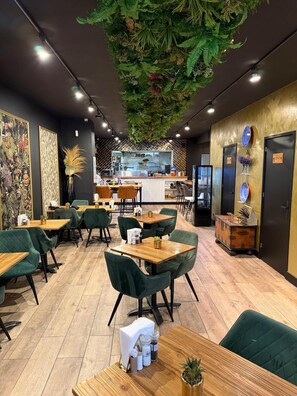 L’intérieur d’un restaurant présente des chaises recouvertes d’un tissu vert, des tables en bois et un espace cuisine central.