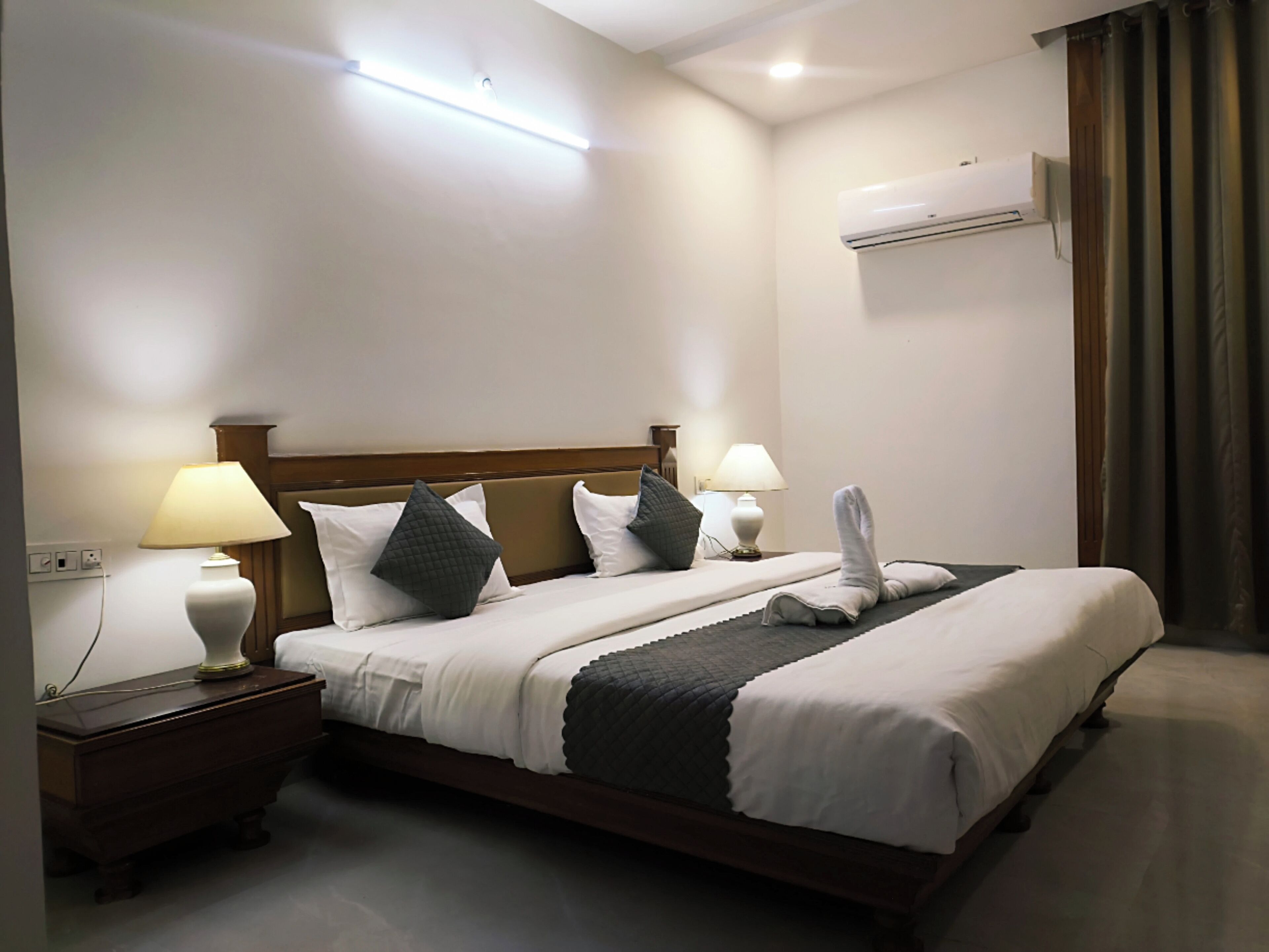 A hotel room with a large bed, two bedside tables with lamps, a wooden headboard, an air conditioning unit, and a curtain.
