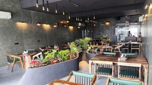 Un interno ristorante moderno con tavoli in legno e sedie imbottite verdi, lampade pendenti a sospensione e diverse piante al centro.