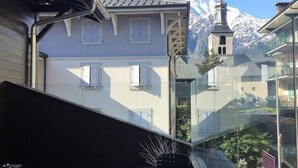 Un balcón con mobiliario exterior, vista a una montaña con la aguja de una iglesia y un edificio moderno con ventanas de vidrio.