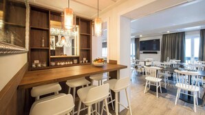 Un bar de hotel moderno con mostrador de madera, taburetes blancos y luces colgantes. Hay varias mesas y sillas en la zona de comedor, y un televisor de pantalla plana montado en la pared.