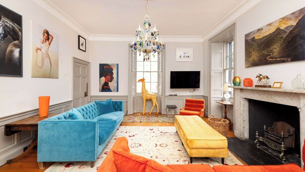 Ein geräumiges Wohnzimmer mit einer großen Couch, einer kleineren Couch, einem gelben Hocker, einem Kamin und einer Chandelier.