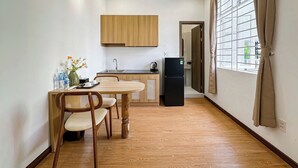 A compact kitchen with wooden cabinets, a dining table with chairs, a refrigerator, and a window with curtains. - The Cozy Nest 1 – Thao Dien (Ho Chi Minh City)