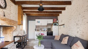 A living room with a grey sofa, a small round table with a vase, a brick fireplace, and a kitchen in the background. - Holiday Home “Le Gîte Aïdan” with Shared Garden and Wi-Fi (LA PUYE)