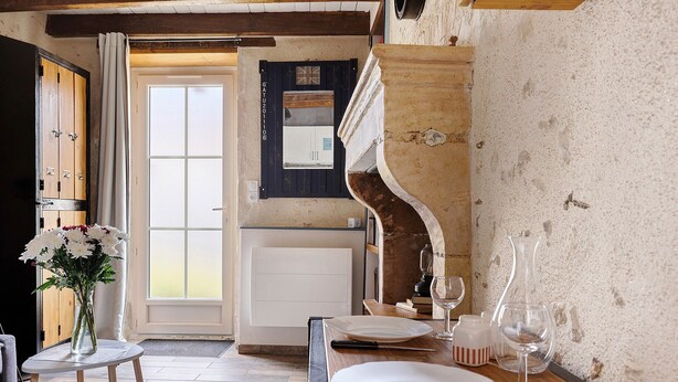 A dining area with a table set for two, a vase with flowers, and a view of the kitchen through an open door. - Holiday Home “Le Gîte Aïdan” with Shared Garden and Wi-Fi (LA PUYE)