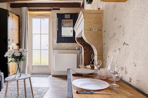 Un coin repas avec une table dressée pour deux personnes, un vase rempli de fleurs et une vue sur la cuisine par une porte ouverte.